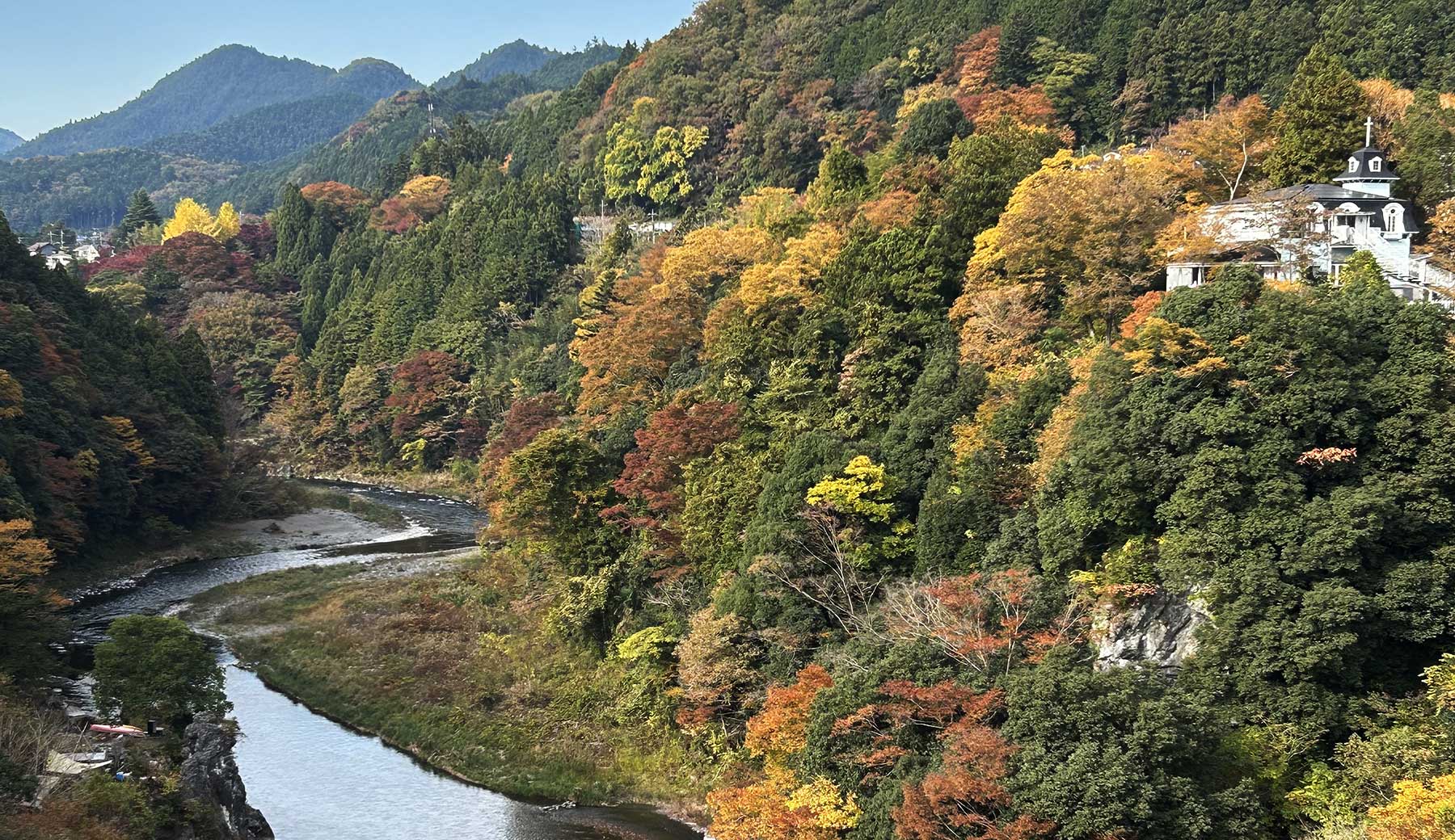 青梅市内の神代橋から眺める紅葉と多摩川の様子。川の両岸の木々が黄色やオレンジに色付いている。