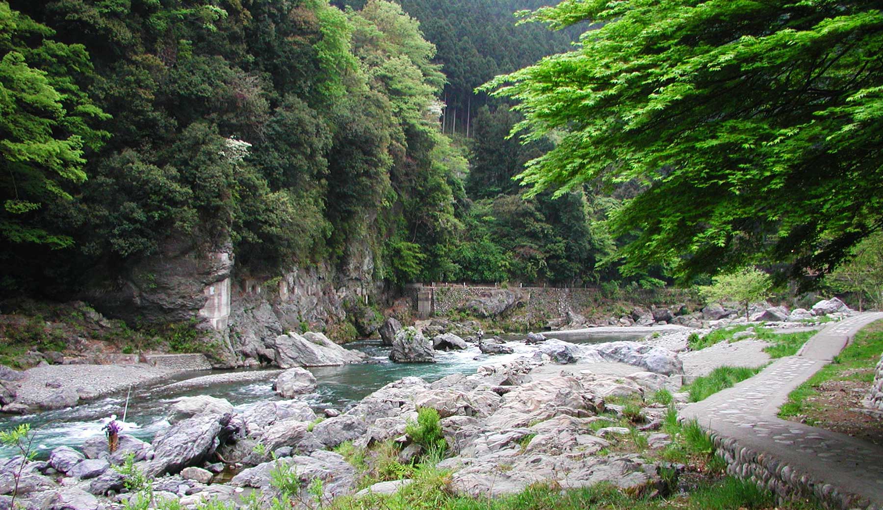 御岳渓谷の遊歩道（御岳園地下流側）の様子。山の手前を多摩川が流れ、河川敷にコンクリートと石で舗装された遊歩道が見える。川の中や岸には人より大きな岩が露出し、その上で釣り人が糸を垂れている。
