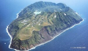 東京都青ヶ島の空撮写真。青い海に断崖に囲まれた緑豊かな島が浮かんでいる。島の中央には巨大なカルデラが見える。