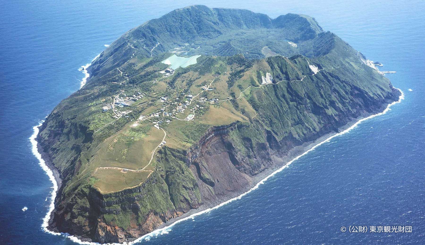 東京都青ヶ島の空撮写真。青い海に断崖に囲まれた緑豊かな島が浮かんでいる。島の中央には巨大なカルデラが見える。