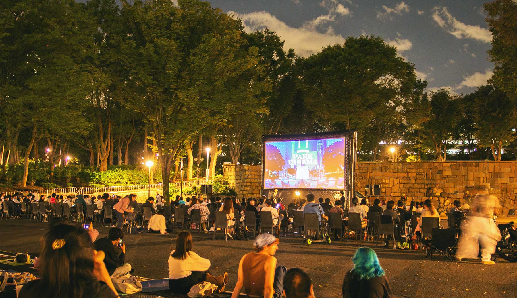 新宿パークシネマフェスティバルの様子。緑豊かな夜の公園にスクリーンが設置され、多くの人が思い思いに野外での映画鑑賞を楽しんでいる。