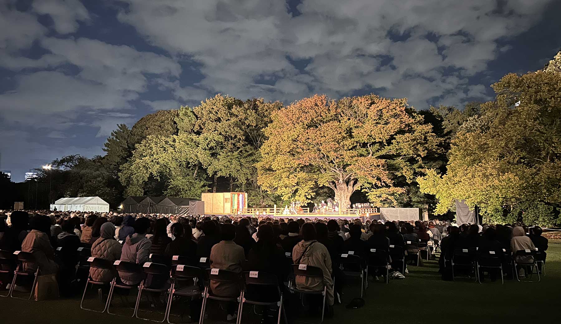「新宿御苑 森の薪能」の様子。新宿御苑の木々を背景に能舞台が置かれ、多数の人々が屋外のに設置された観覧席で薪能を鑑賞している。