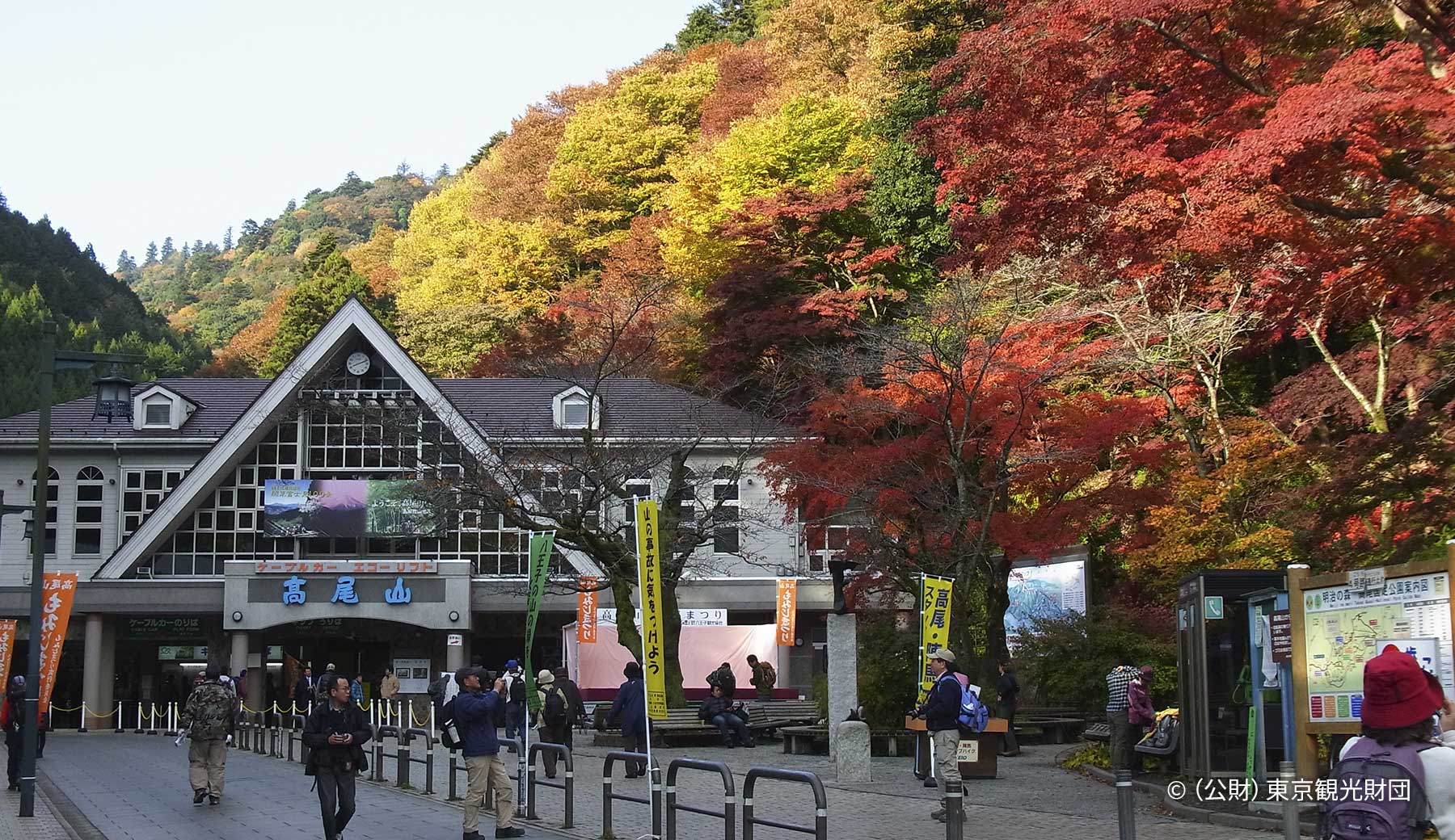 高尾山の麓にあるケーブルカー駅「清滝駅」駅前広場の秋の様子。三角屋根が特徴の駅舎周囲は色とりどりの紅葉に覆われ、大勢の登山客が写真を撮ったり休憩したりしている。
