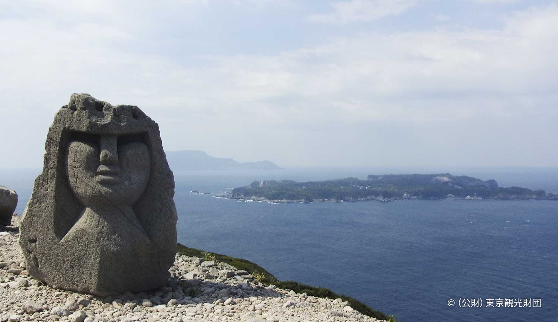 新島の石山展望台にある大きなモヤイ像の写真。周辺の島々が浮かぶ海を背景に、人物の方から上を模した石像が立っている。